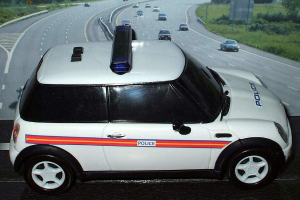 Ein weißes Polizeifahrzeug steht im Vordergrund, dahinter sind mehrere Autos auf der Straße unterwegs, umgeben von Gras und Bäumen auf beiden Seiten.