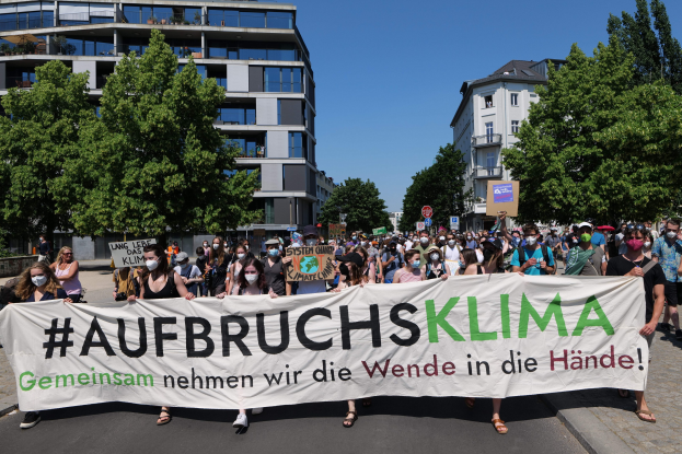Menschen mit Masken halten ein Transparent mit der Aufschrift 'Aufbruchsklima' vor einem Gebäude mit Bäumen und einem klaren Himmel.