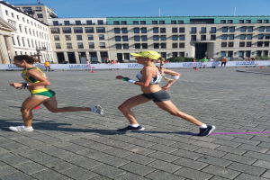 Zwei Frauen in Sportkleidung laufen bei einem Marathon auf einer Stadtstraße, umgeben von Zuschauern, einem Zaun, einer Fahne, Pfosten, Pflanzen, einem Gebäude mit Fenstern und einem bewölkten Himmel.