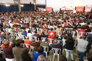 Ein volles Stadion mit vielen sitzenden und stehenden Menschen, eine Bühne mit zwei Rednern an einem Podium und mehreren Fahnen und Lichtern in der Nähe.