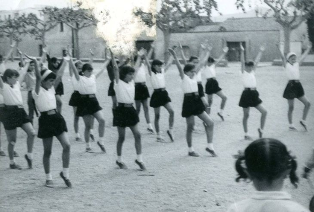 Schwarzes und weißes Foto von jungen Mädchen in Schuluniformen, die auf einer von Gebäuden und Bäumen gesäumten Straße tanzen, mit einer Person in Schuhen unten.
