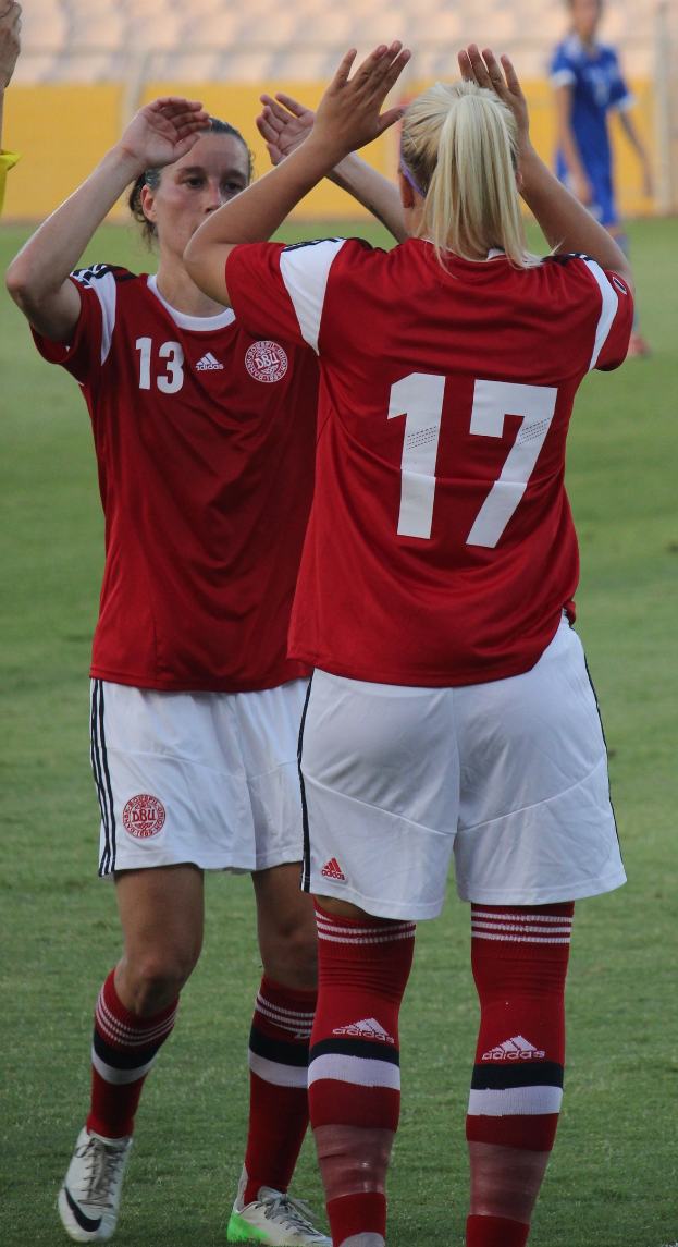 Zwei Frauen in roten Hemden und weißen Shorts klatschen auf einem Fußballfeld, mit einer Person in einem blauen Kleid und einer gelben Tafel im Hintergrund.