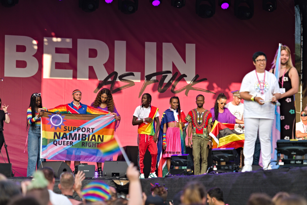 Menschen auf einer Bühne mit einem Banner "Berlin as Fuck" und Lautsprechern, Mikrofonen und einer Menge unten mit hochgereckten Händen.