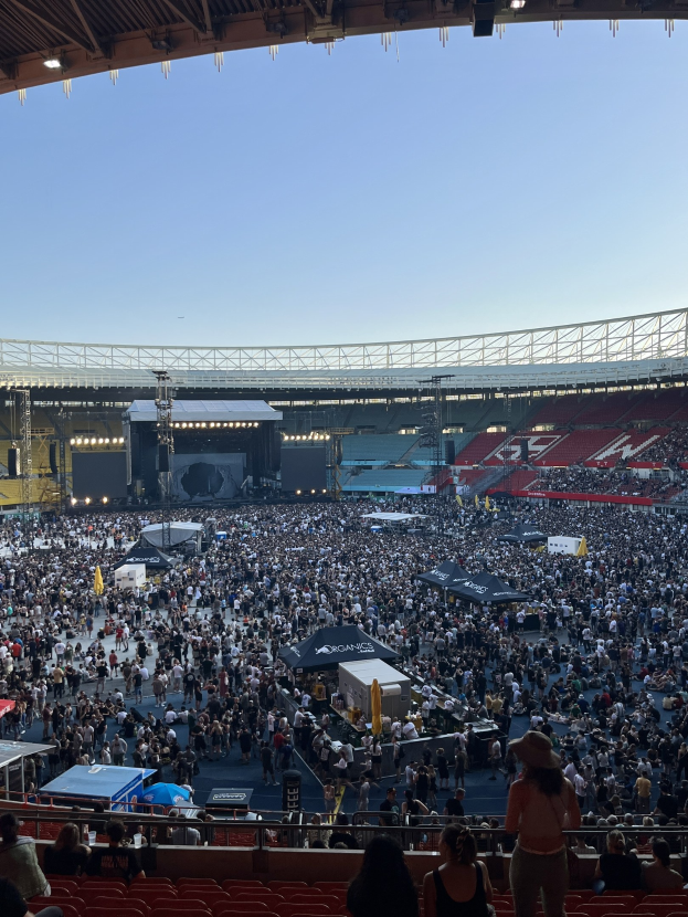 Eine große Menschenmenge sitzt und steht in einem Stadion mit Geländern, Zelten und Lichtern unter einer Decke mit Überkopfbeleuchtung vor einem Himmel als Hintergrund.