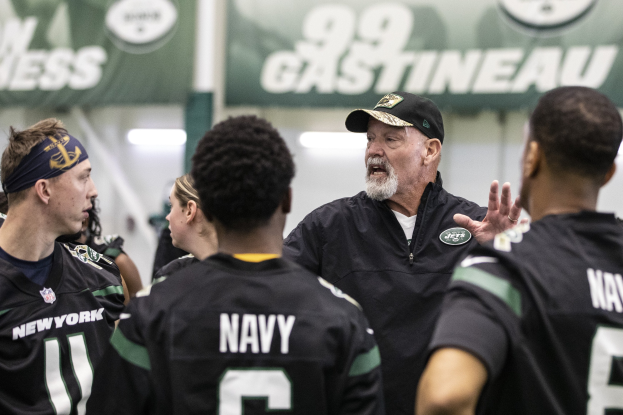 Gruppe von Männern in einem Stadion, mit Jets-Headcoach Mike McCarthy in einer Mütze, der zu seinem Team spricht, umgeben von Bannern und Lichtern.
