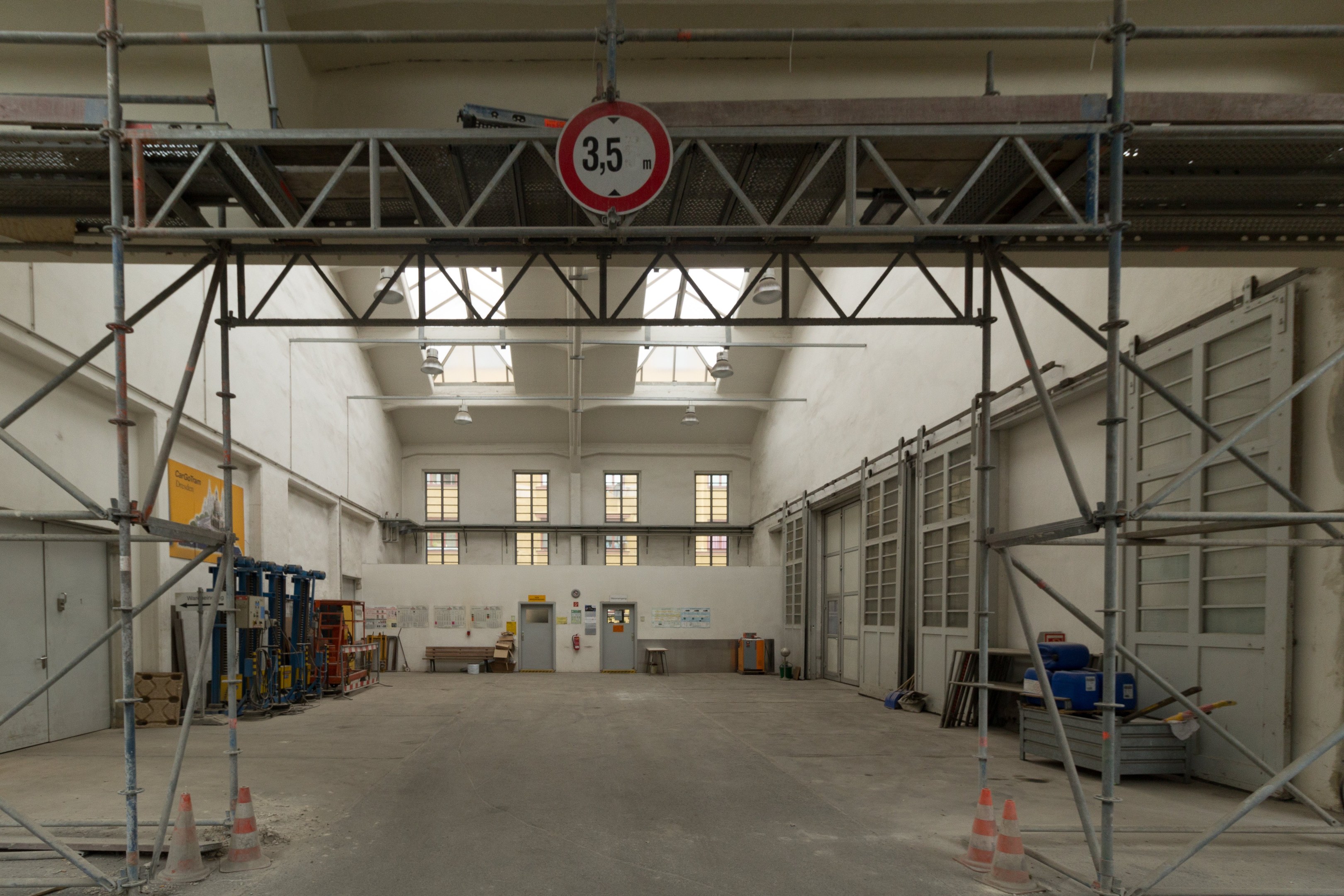Lagerhalle mit Gerüsten, Verkehrskegeln, Metallstangen, einem Schild, Maschinen, einer Tür, Fenstern, einer Wand, Deckenleuchten und Dach.