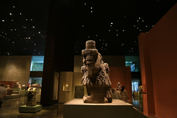 Eine Gruppe von Menschen in einem Museum, die Skulpturen auf Sockeln unter Deckenlampen betrachtet, mit Museumswänden im Hintergrund.