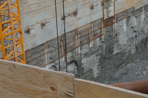 Baustelle mit einem Kran auf der linken Seite, einer Betonwand im Hintergrund, einem hölzernen Objekt und einem Rohr unten.