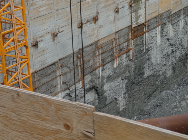 Baustelle mit einem Kran auf der linken Seite, einer Betonwand im Hintergrund, einem hölzernen Objekt und einem Rohr unten.