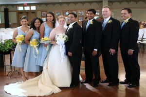 Eine Frau in einem weißen Kleid mit einem Bouquet steht in der Mitte, umgeben von Frauen in blauen Kleidern mit Bouquets und Männern in schwarzen Anzügen, die alle lächeln und drinnen mit verschiedenen Gegenständen im Hintergrund stehen.