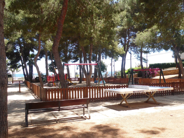 Eine Parkszene mit einem zentralen Tischtennis-Tisch, umgeben von Bänken, Schaukeln und Grünflächen, mit einem Zaun, Pfosten und Bäumen, vor einem Gewässer und einem klaren blauen Himmel.