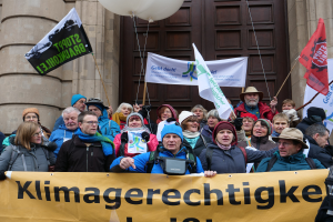 Eine Gruppe von Menschen vor einem Gebäude mit Säulen und einer Tür, die eine Fahne mit Text, Fahnenstangen und Luftballons halten, nimmt an einem Klimastreik in Deutschland teil.