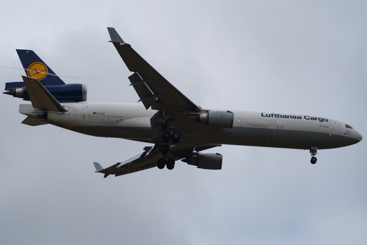 Lufthansa Cargo Boeing 777-300ER im Flug mit sichtbarem Text auf dem Rumpf, ausgefahrenen Flügeln, aktiven Triebwerken, gegen einen Himmel mit weißen Wolken.