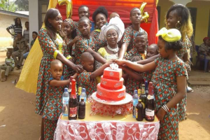 Eine Gruppe von Menschen steht um einen Tisch mit einem Kuchen, Flaschen und anderen Gegenständen in einer ländlichen Umgebung mit Häusern, Bäumen und einem klaren blauen Himmel, feiert einen Geburtstag mit einigen, die sich an den Händen halten und andere, die lächeln.