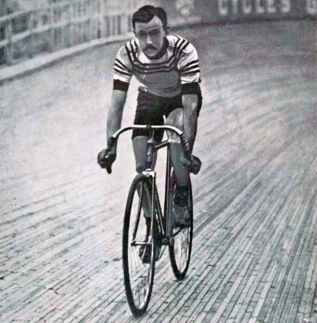 Schwarz-weiß-Foto eines Mannes, der auf einer Bahnradsportanlage fährt, mit einem Zaun und einer Tafel im Hintergrund.