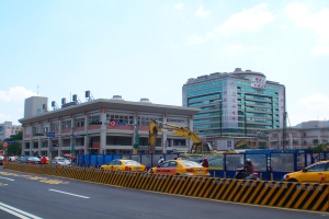 Eine belebte Stadtstraße mit starkem Verkehr, hohen Gebäuden, Fußgängern, einem Kran, Verkehrsampeln, Schildern, Bäumen und einem bewölkten Himmel.
