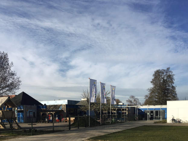 Großes Schulgebäude mit Metallzaun, Rasenfläche, Weg, Bäumen, Schildern, einem geparkten Fahrrad, nahen Häusern und einem bewölkten Himmel.