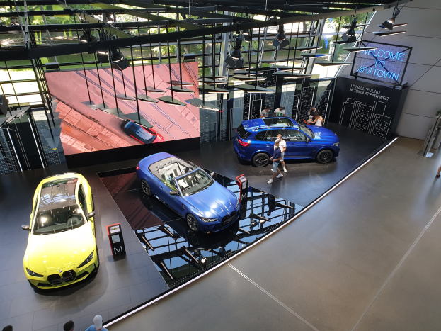 Eine Gruppe von Menschen, die im BMW Museum in München stehen, mit Autos auf der linken Seite und einem Schild mit der Aufschrift "BMW Museum München" auf der rechten Seite, mit Blick auf eine Außenszene von Böumen, Geböuden und Himmel durch Glasfenster.