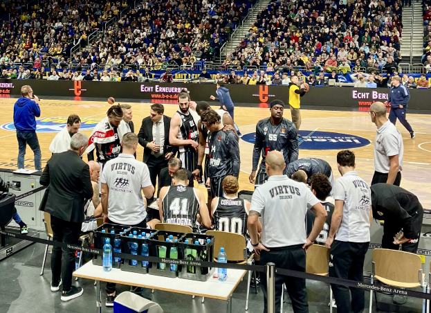 Eine Gruppe von Männern auf einem Basketballfeld mit einer Menge, die um sie herum sitzt und steht, ein Fotograf mit Ausrüstung auf einem Tisch links und Hintergrundtafeln mit den Teamnamen "Virtus Berlin" und "Echt Berlin."