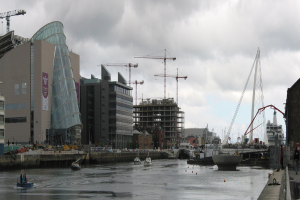 Ein Fluss mit Schiffen, eine Brücke, Gebäude, Baukräne und der Himmel im Hintergrund.