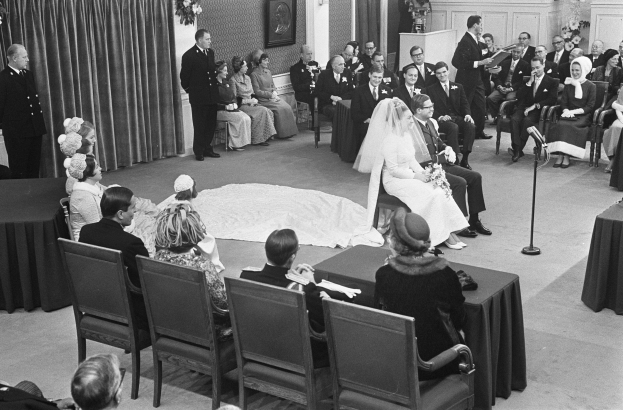 Schwarzes und weißes Foto einer Hochzeitszeremonie mit sitzenden und stehenden Gästen, einem Mann, der in ein Mikrofon spricht, einer Frau mit einem Blumenstrauß, einem Podium mit einem Mikrofon, Blumengestecken, einem Vorhang, einem Rahmenfoto an der Wand und einer Tür im Hintergrund.