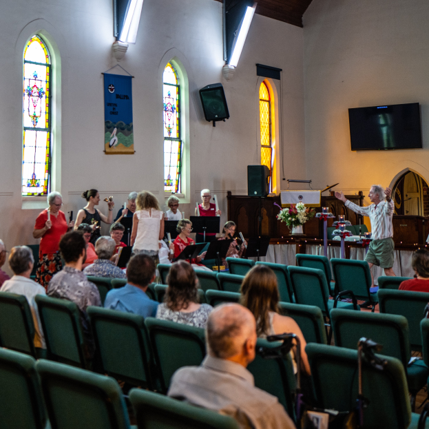 Eine Gruppe von Menschen sitzt auf Stühlen in einer Kirche, ein Mann steht vorne mit einem Mikrofon, umgeben von Musikinstrumenten, einem Tisch mit einer Blumenvase, einem Lautsprecher, einer Banner mit Text, Fenstern, einem Fernseher und Deckenleuchten.