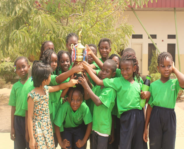 Gruppe von Kindern mit einem Pokal in einem Schulhof mit Bäumen und einem Gebäude im Hintergrund.