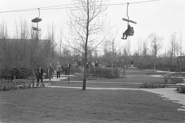 Schwarzes und weißes Foto von Menschen, die eine Skilift in einem Park mit Bäumen, Pflanzen, Gras und einem Weg fahren, mit Gebäuden und einem klaren Himmel im Hintergrund.