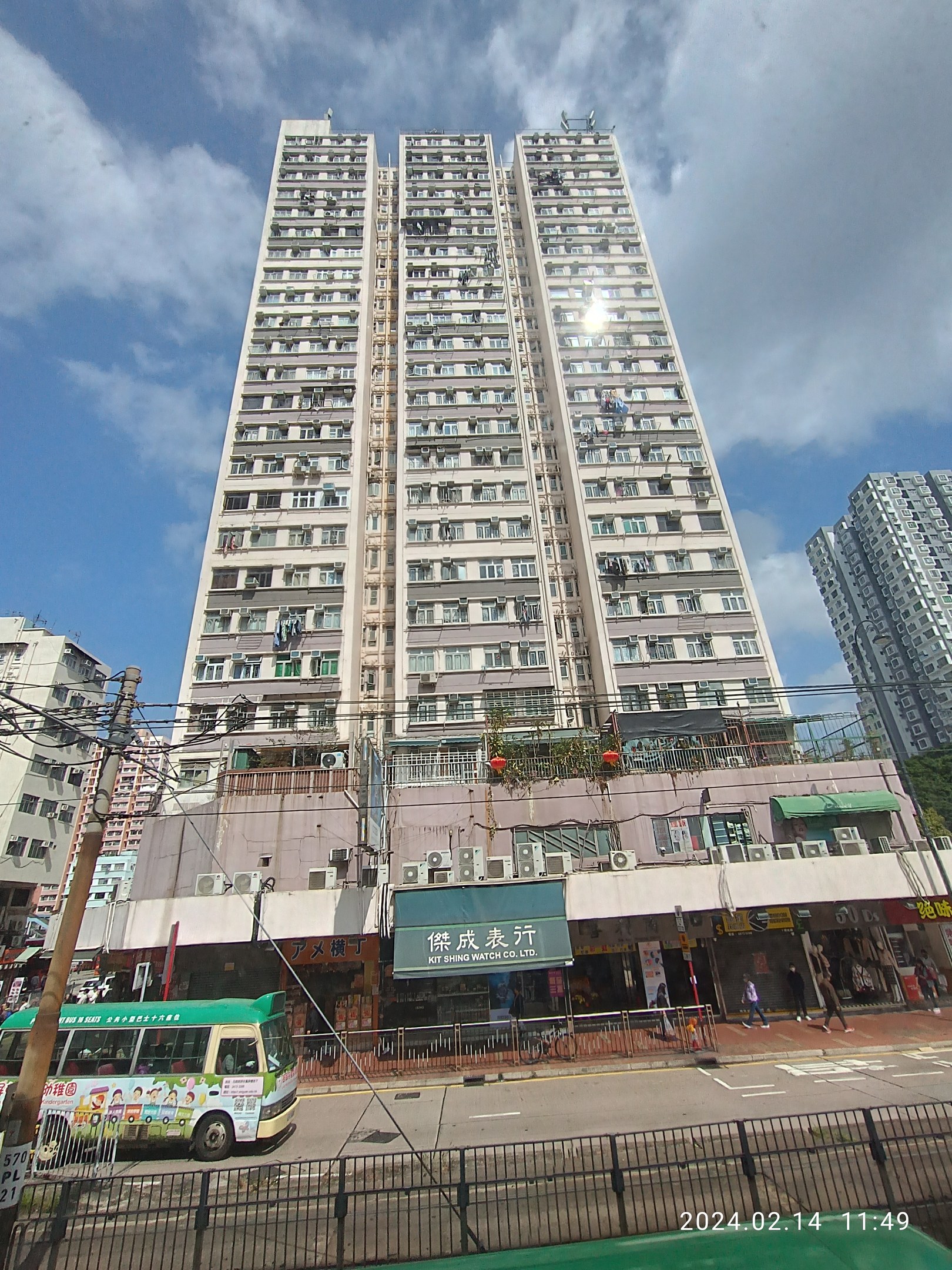 Ein hoher Wohnblock auf einer belebten Stadtstraße umgeben von anderen Gebäuden, Fahrzeugen, Fußgängern und urbanen Elementen unter einem bewölkten Himmel.