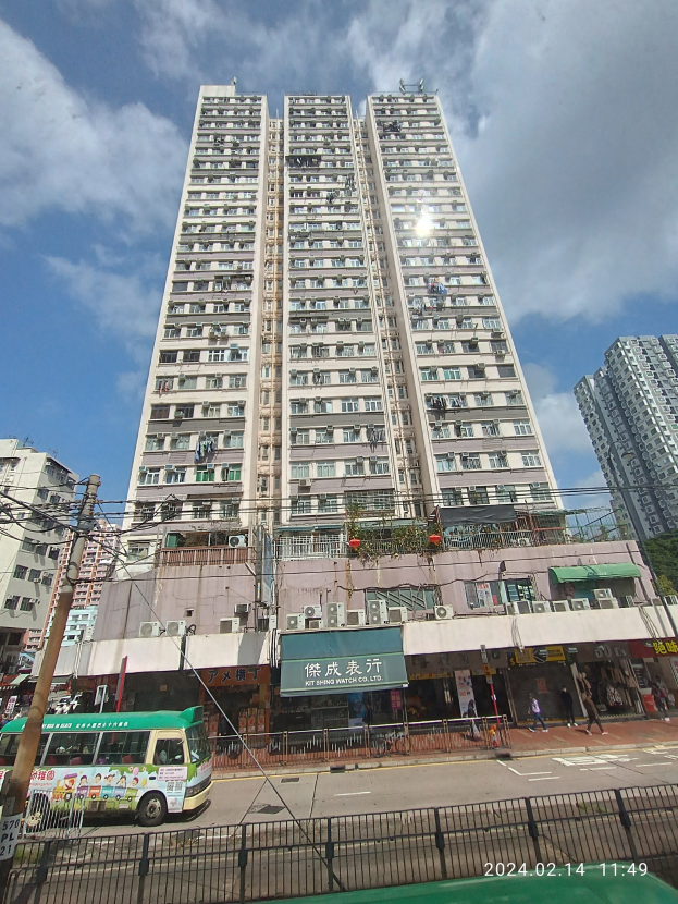 Ein hoher Wohnblock auf einer belebten Stadtstraße umgeben von anderen Gebäuden, Fahrzeugen, Fußgängern und urbanen Elementen unter einem bewölkten Himmel.