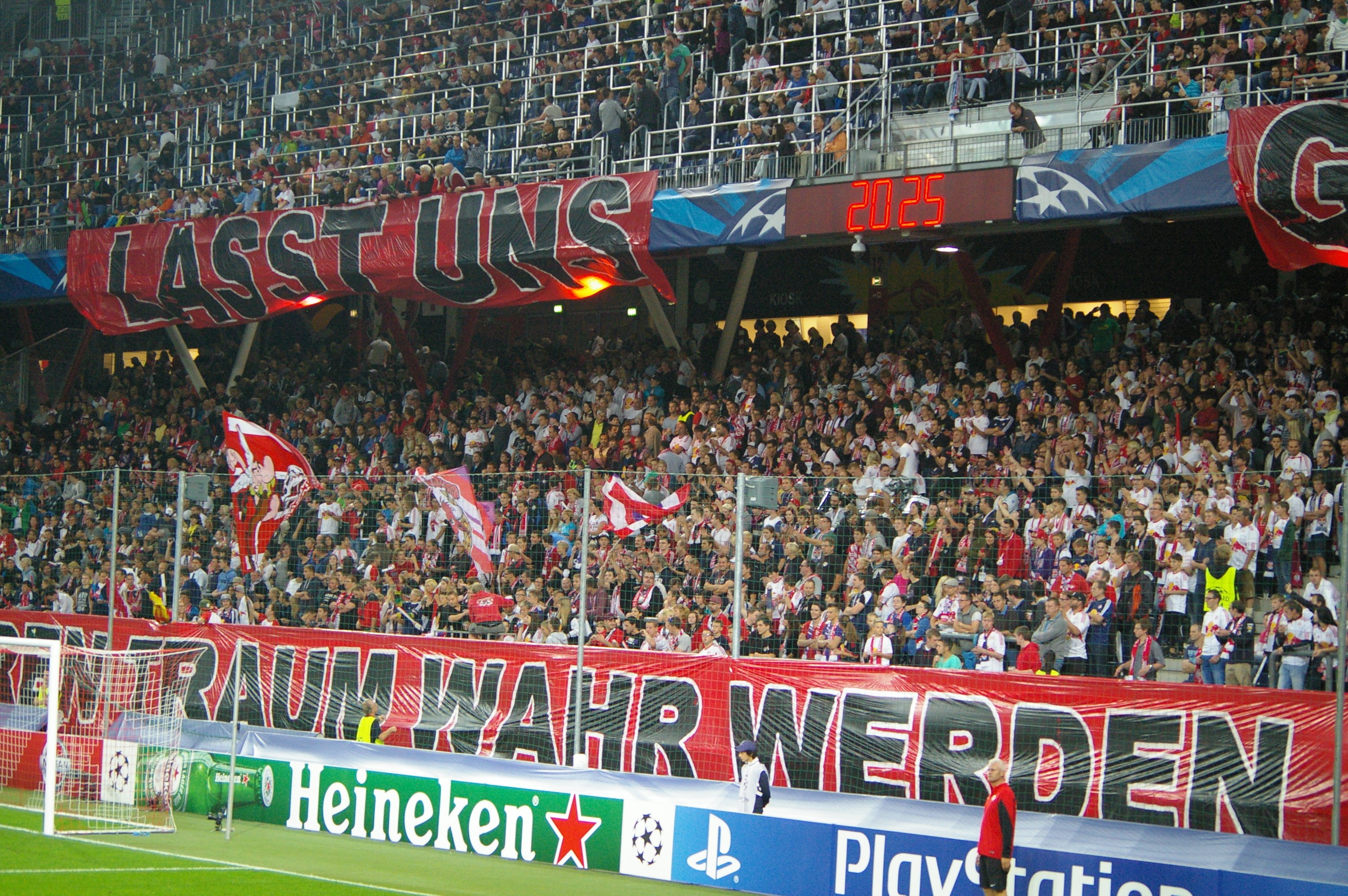 Fußballspiel in einem Stadion mit einer großen Zuschauermenge, saftigem Grün, Torpfosten links, Bodenbanner, Menschen mit Fahnen und Hintergrundgeländer, Tafeln, Lichter und Gegenstände.