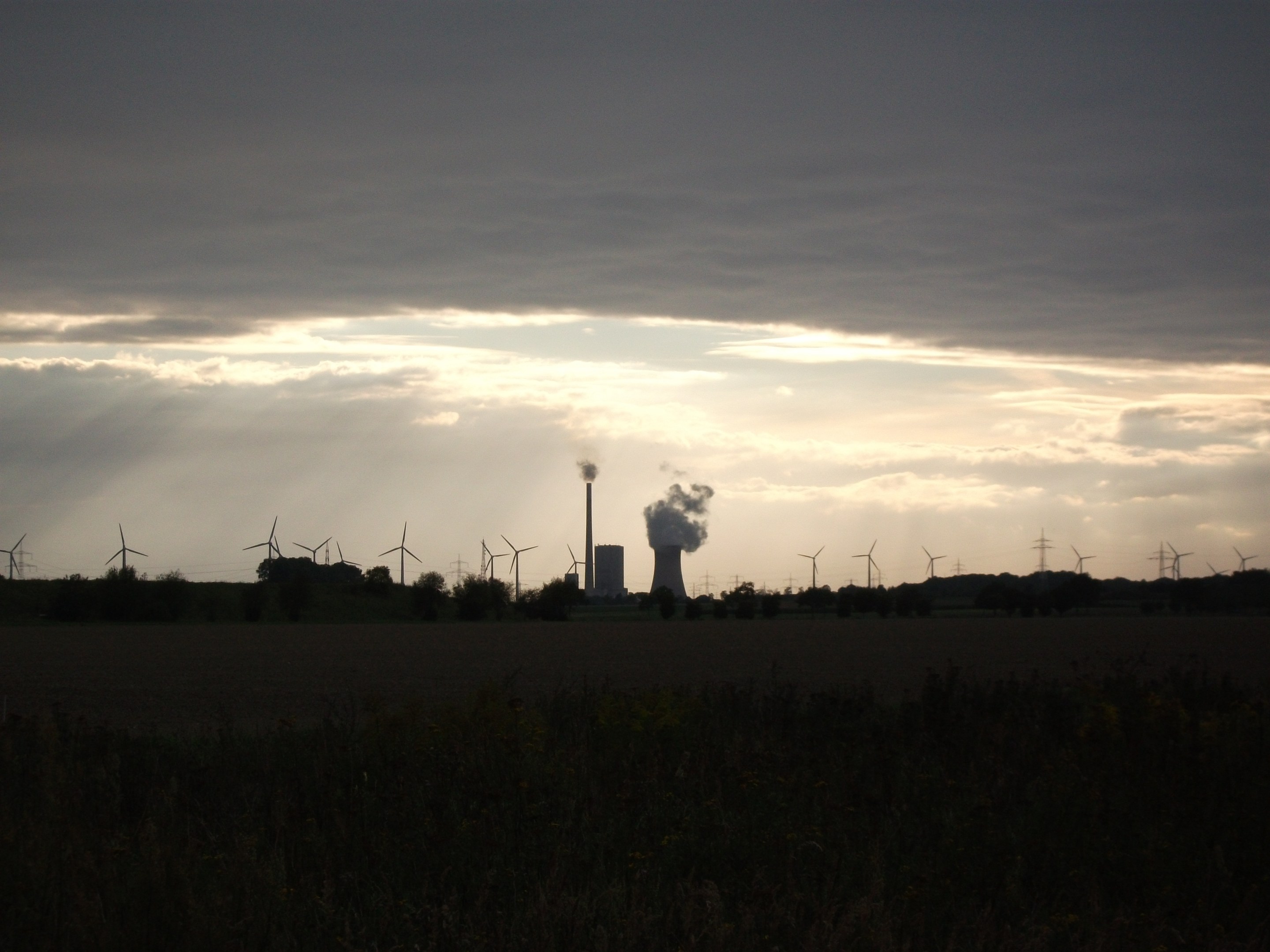 Kraftwerk mit Windrädern vor wolkenbedecktem Himmel, umgeben von Grün am Boden.