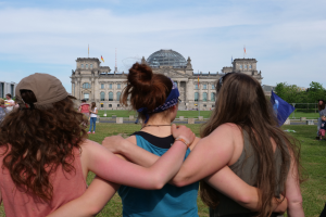 Drei Frauen in verschiedenen farbigen Kleidern stehen vor dem Reichstaggebäude in Berlin, Deutschland, mit Menschen, Flaggen, Bäumen und Wolken im Hintergrund.