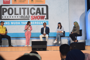 Eine Gruppe von Menschen sitzt auf einer Bühne mit einer Frau in der Mitte, die ein Mikrofon hält, einer Zuschauermenge im Vordergrund und einem Banner im Hintergrund mit der Aufschrift 'Politische Straßen-show 2019'.