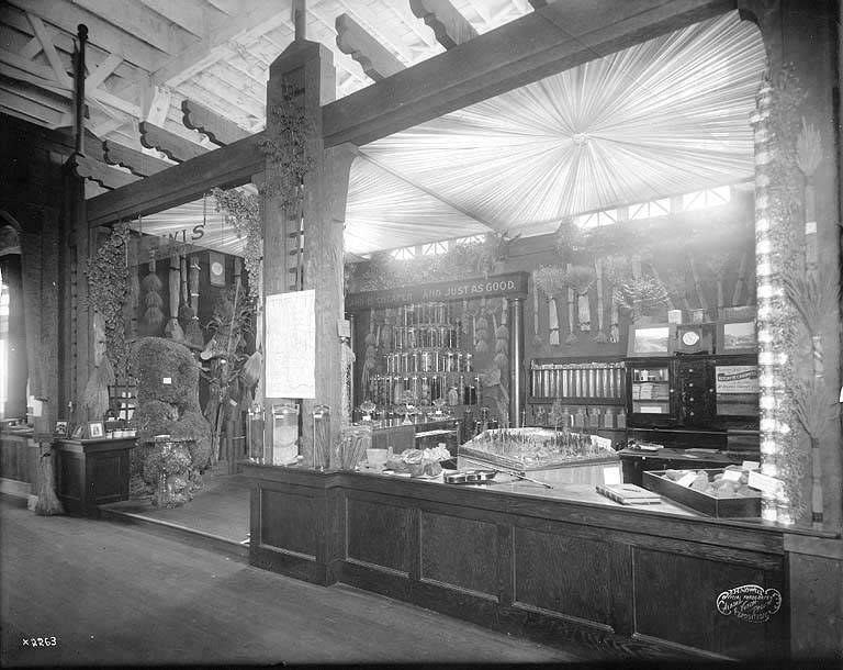 Altes Schwarz-Weiß-Foto einer Apotheke mit einem zentralen Tresen, auf dem verschiedene Gegenstände stehen, Regalen im Hintergrund mit Flaschen und Artikeln gefüllt und Text unten, der darauf hinweist, dass es sich um eine Apotheke handelt.