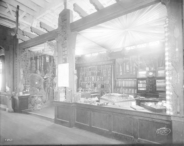 Altes Schwarz-Weiß-Foto einer Apotheke mit einem zentralen Tresen, auf dem verschiedene Gegenstände stehen, Regalen im Hintergrund mit Flaschen und Artikeln gefüllt und Text unten, der darauf hinweist, dass es sich um eine Apotheke handelt.