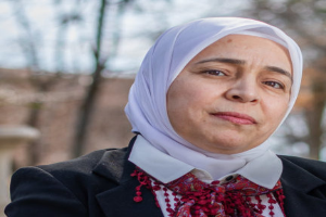 Juli Tymoshenko, mit einem weißen Kopftuch und einer roten Schärpe bekleidet, steht vor einem unscharfen Hintergrund aus Bäumen und einem Gebäude.