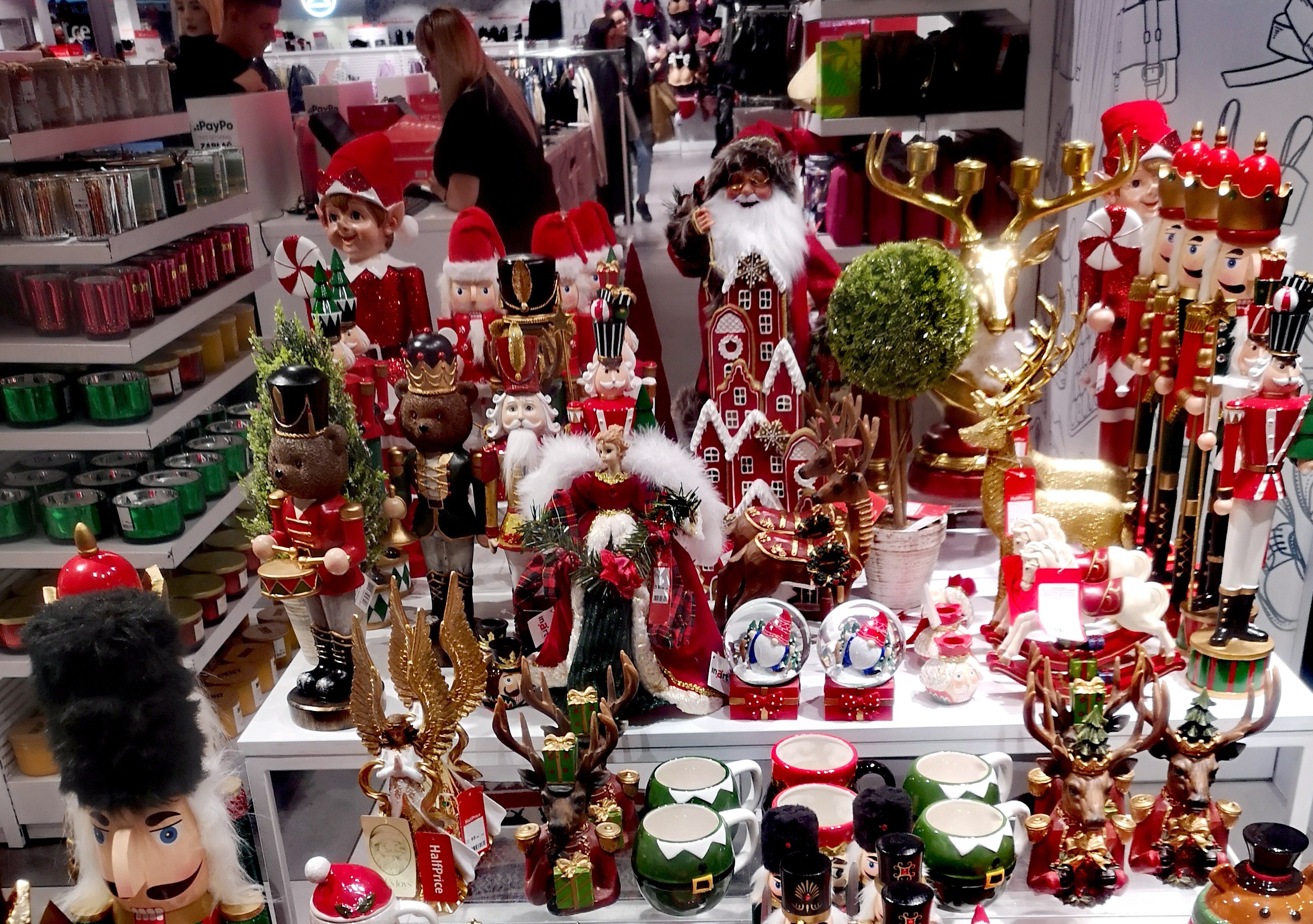 Ladeninneres mit Weihnachtsdekorationen, darunter Spielzeug, Tassen und andere Gegenstände auf Tischen und Regalen, mit Menschen, Lichtern und einer Wand im Hintergrund.