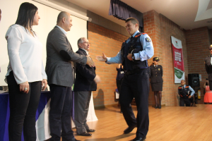 Eine Gruppe von Menschen steht auf einer Bühne mit einer uniformierten Person, die eine Mütze hält, einer Frau in einem weißen Hemd, einem Banner, einem Lautsprecher auf einem Ständer, Vorhängen, einem Tisch und einem Wandbildschirm im Hintergrund.