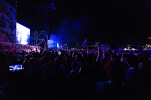 Eine Menschenmenge bei einem Konzert mit erhobenen Händen, einige halten Mobiltelefone und Fahnen, mit einem Anzeigebildschirm, Metallrahmen und Lichtern im Hintergrund.