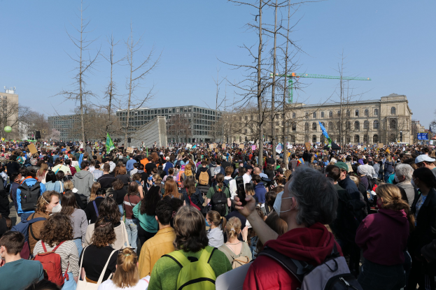 Eine große Menge von Menschen, die an einer Klimaprotestaktion vor einem Gebäude mit Fenstern, Bäumen und einem klaren Himmel teilnehmen, viele halten Schilder und tragen Taschen.