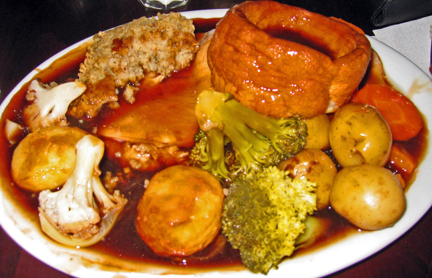 Ein weißer Teller mit Yorkshire-Pudding und verschiedenen Gemüsen wie Karotten, Kartoffeln und Zwiebeln, das auf einem Tisch mit Gläsern, Papiertüchern und anderen Gegenständen drumherum steht.