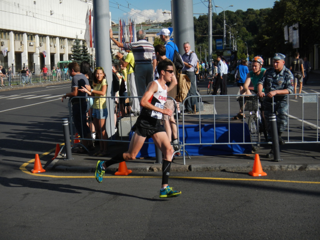 Ein Mann läuft bei einem Marathon auf einer Stadtstraße, umgeben von Zuschauern auf dem Gehweg, einige halten Fahrräder, mit städtischer Infrastruktur und einem bewölkten Himmel im Hintergrund.