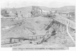 Ein Schwarz-Weiß-Foto eines Kohlebergwerks in Kolumbien, das ein Rad im Vordergrund, Gebäude, Pfosten, Bäume und Berge im Hintergrund zeigt, mit Text am unteren Bildrand.