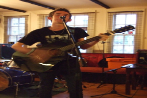 Ein Mann in schwarzem T-Shirt und Hose steht mit einer Gitarre und einem Mikrofon auf einem Ständer, mit einer blauen Trommel in der linken Ecke, einer Couch mit Gegenständen darauf und durchsichtigen Vorhängen im Hintergrund unter einer Decke.