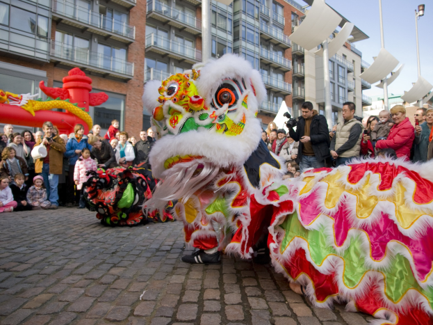 Ein lebendiges chinesisches Neujahrsfest in Amsterdam mit einer Löwen-Tanzvorstellung vor einer Zuschauermenge, darunter einige mit Kameras, vor einem Hintergrund aus Gebäuden, Laternenmasten und einem klaren blauen Himmel.