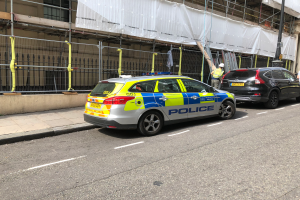 Ein Polizeiwagen steht auf der Straße neben einem Gebäude, daneben stehen zwei Helme tragende Personen; im Vordergrund sind ein Geländer und ein Pfahl zu sehen, im Hintergrund hängt ein weißes Tuch über das Gebäude.