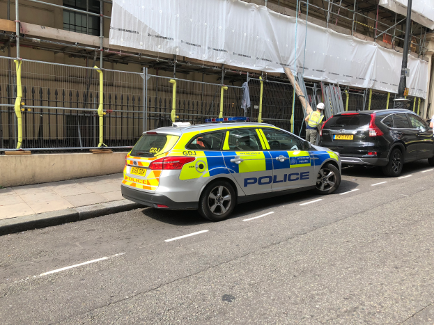 Ein Polizeiwagen steht auf der Straße neben einem Gebäude, daneben stehen zwei Helme tragende Personen; im Vordergrund sind ein Geländer und ein Pfahl zu sehen, im Hintergrund hängt ein weißes Tuch über das Gebäude.