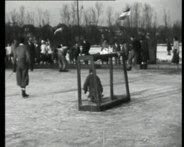 Schwarzes und weißes Foto einer Gruppe von Menschen, die Hockey auf einem Eisplatz spielen, mit einem Netz in der Mitte, Bäumen, Pfosten und einer Fahne im Hintergrund und Text oben und unten.