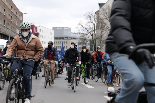 Eine Gruppe von Radfahrern in Helmen und Handschuhen fährt eine von Bäumen gesäumte Straße in Berlin entlang, mit Gebäuden und geparkten Fahrzeugen im Hintergrund und einem klaren Himmel.